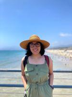 A picture of Sofía, smiling, wearing a wide-brimmed straw hat, thick-rimmed glasses, a red backpack, and jade green dress, leaning against the blue railing of an elevated platform with crystal-clear ocean water meeting a sandy beach below.