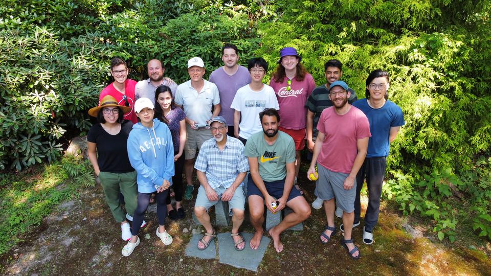 A picture of the Aziz group posed in two rows in Mike's backyard. Behind the group is lush, green foliage from several bushes. Mike and Thomas are seated in the center of the group on a stone bench.