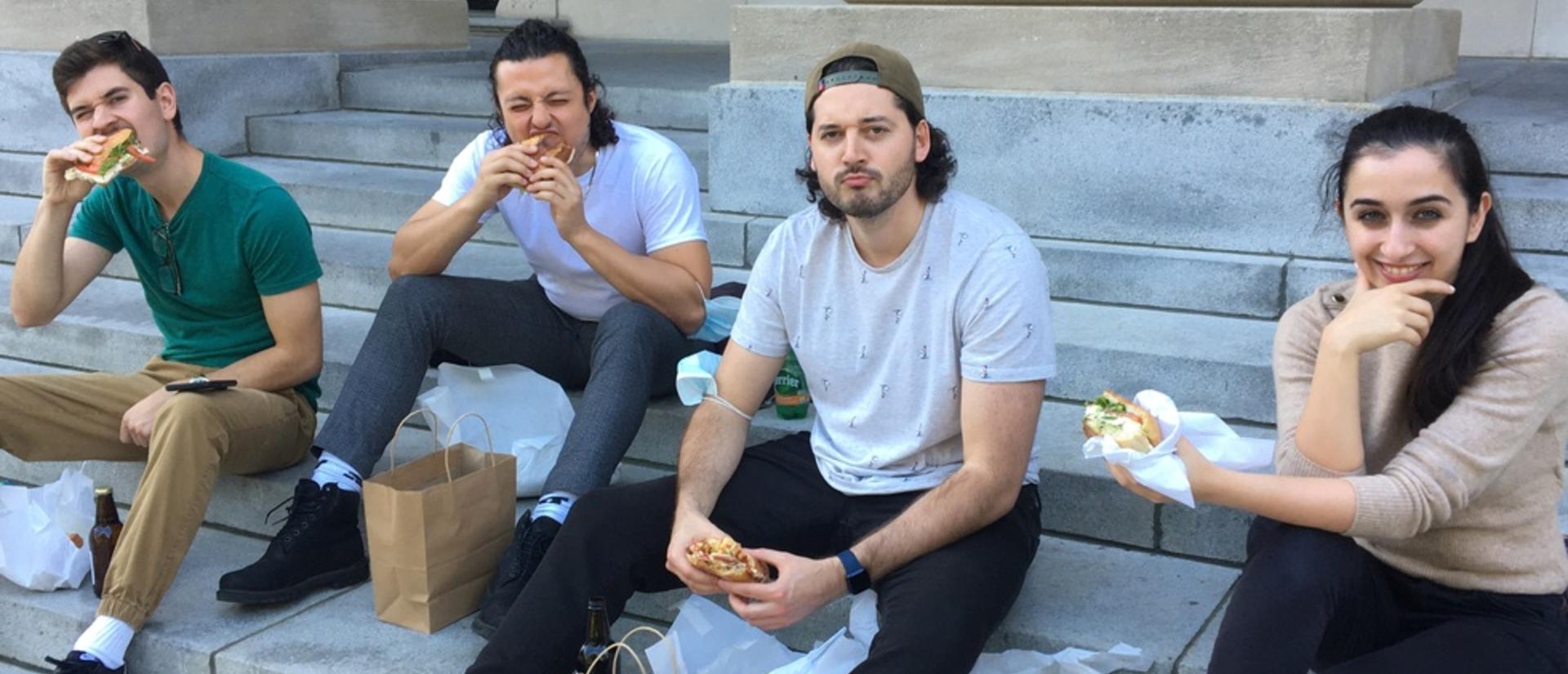 Kiana, Eric, Anton, and Jordan sitting on stone library stairs, enjoying sandwiches