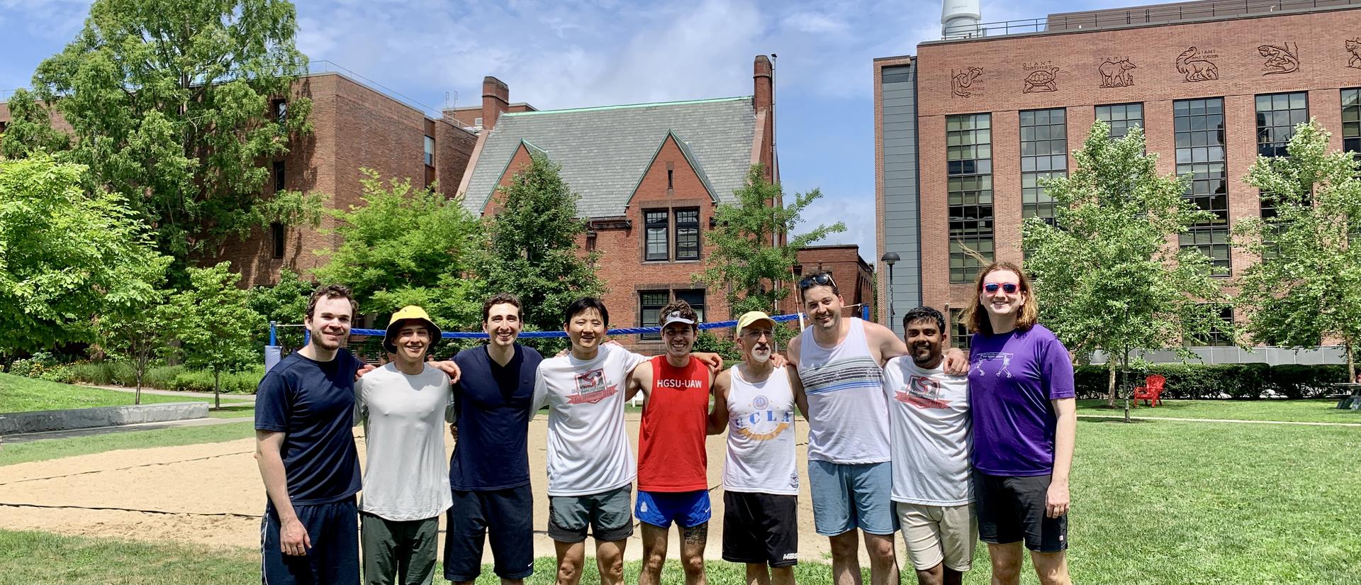 The volleyball team stands on grass in the courtyard of the biology labs, with a sandy beach volleyball court behind.
