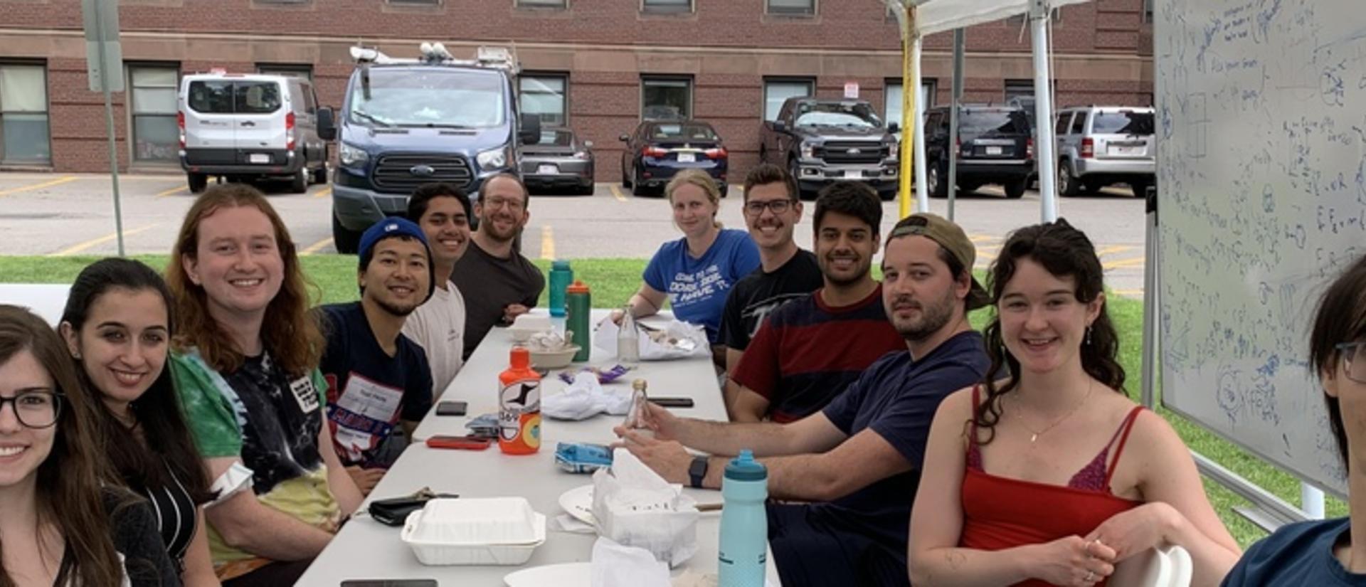 Aziz group and friends sit at plastic picnic tables under a white pavilion, gathered for summer lunchtime