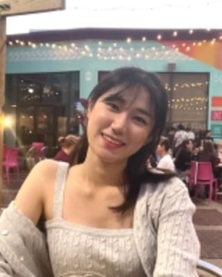 Kyumin is smiling, wearing a beige sweater, seated on a patio where others are sitting in magenta chairs at tables in groups, all in front of a teal building with large windows, draped with strings of lights.