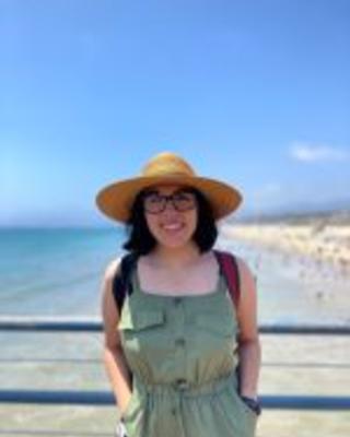 A picture of Sofía, smiling, wearing a wide-brimmed straw hat, thick-rimmed glasses, a red backpack, and jade green dress, leaning against the blue railing of an elevated platform with crystal-clear ocean water meeting a sandy beach below.