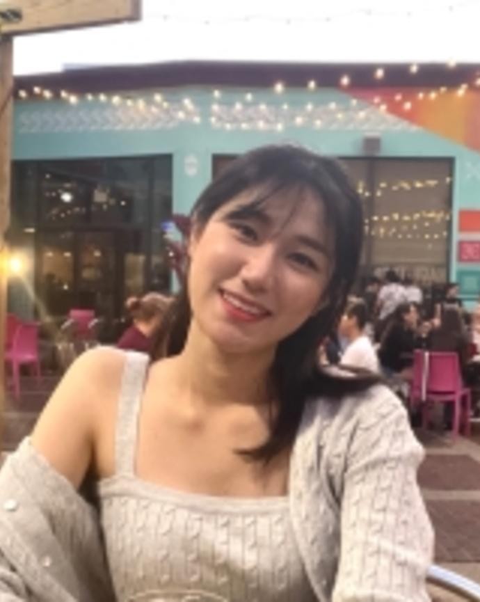 Kyumin is smiling, wearing a beige sweater, seated on a patio where others are sitting in magenta chairs at tables in groups, all in front of a teal building with large windows, draped with strings of lights.