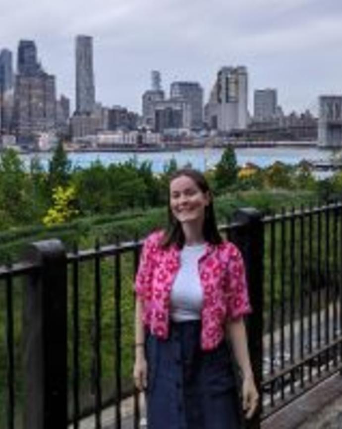 Sara is standing in front of a black painted metal fence, which is in front of some greenery and a body of water. A city stands on the other side of the water, skyscrapers reaching to the grey clouds.
