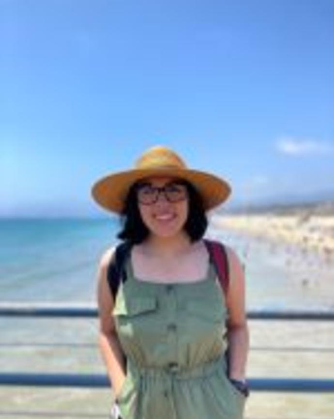 A picture of Sofía, smiling, wearing a wide-brimmed straw hat, thick-rimmed glasses, a red backpack, and jade green dress, leaning against the blue railing of an elevated platform with crystal-clear ocean water meeting a sandy beach below.