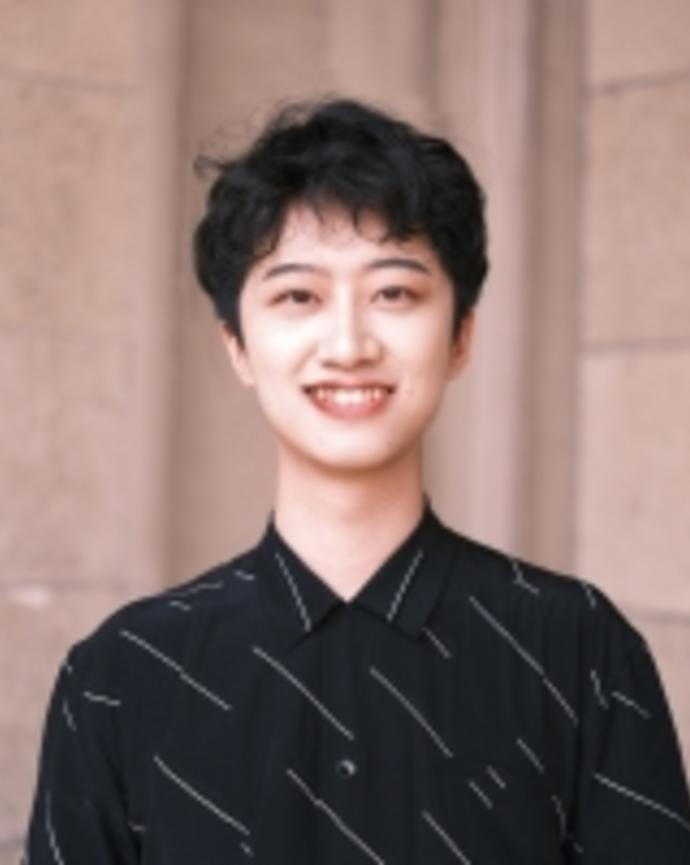 A picture of Taobo, smiling, wearing a black collared shirt with a few thin white stripes, stinding in front of a background of a sandy-colored stone building.