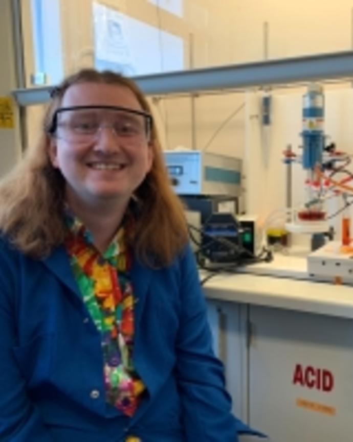 Tommy is sitting next to a fume hood, wearing a blue labcoat over a rainbow floral patterned shirt
